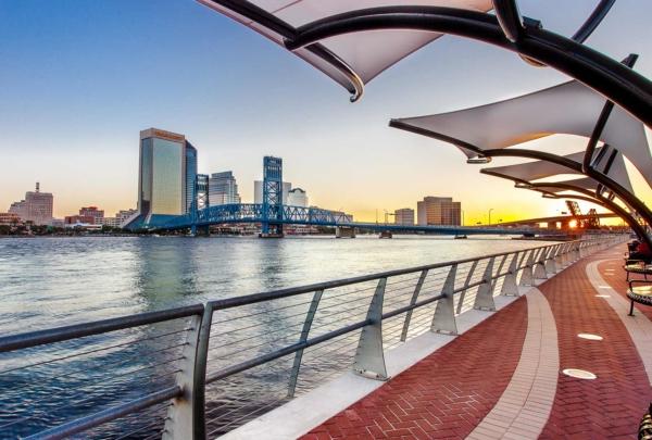 Photo of Jacksonville's Southbank Riverwalk at sunset. 河边的红砖人行道上有钢护栏. Canopies overhang the sidewalks. Jacksonville skyline in background.